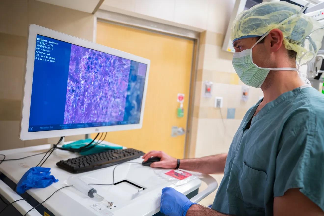 Dr Todd Hollon wears teal scrubs, a mask, and hairnet while standing at a computer with a medical image on the screen