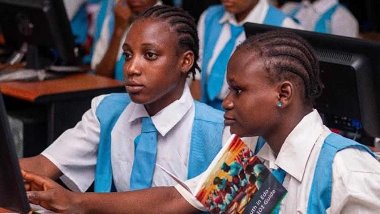 Two black students with cornrows wearing white shirts and blue vests and ties look at a computer