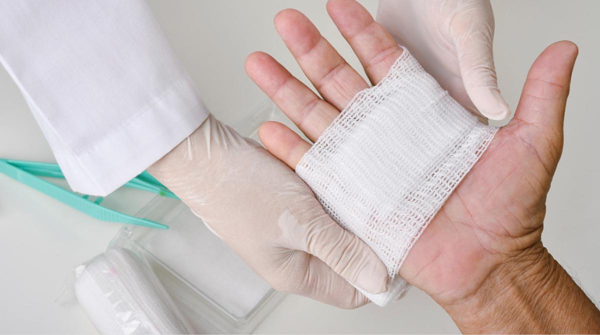 An outstretched hand with a white bandage around it. The bandage is being applied by hands wearing medical gloves and a white lab coat. 