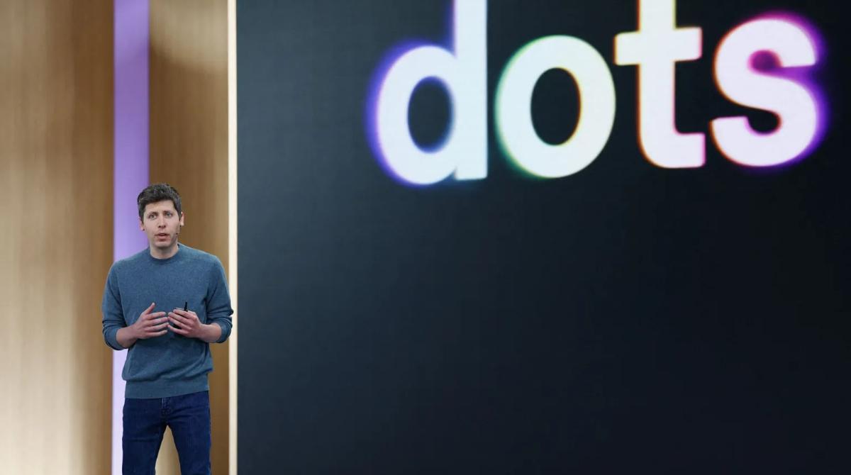 Sam Altman stands to the left of a screen showing the word "dots". 