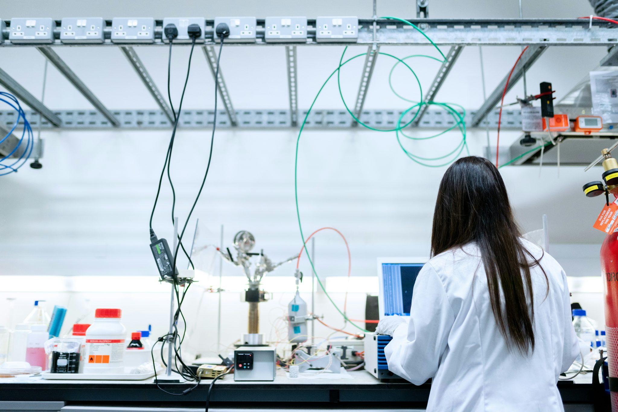  A scientist in a white lab coat works at a computer in a laboratory surrounded by scientific equipment, bottles, cables, and tubing. The researcher is shown from behind at a busy lab bench, with power outlets and experimental apparatus visible throughout the workspace.